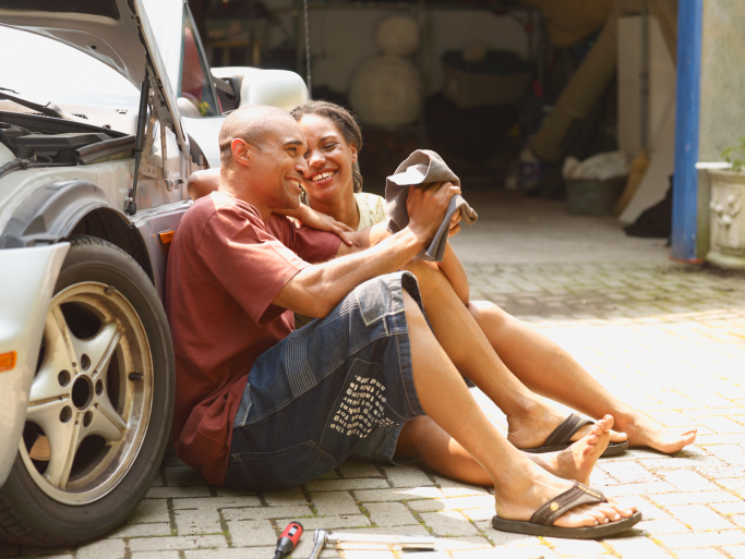 Couple sitting on the ground outside garage next to car laughing