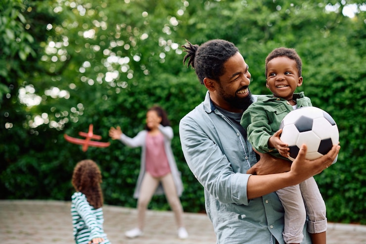 Father holding his son and a soccer ball in the foreground and others playing with a toy plane in the background