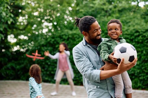 Father holding his son and a soccer ball in the foreground and others playing with a toy plane in the background