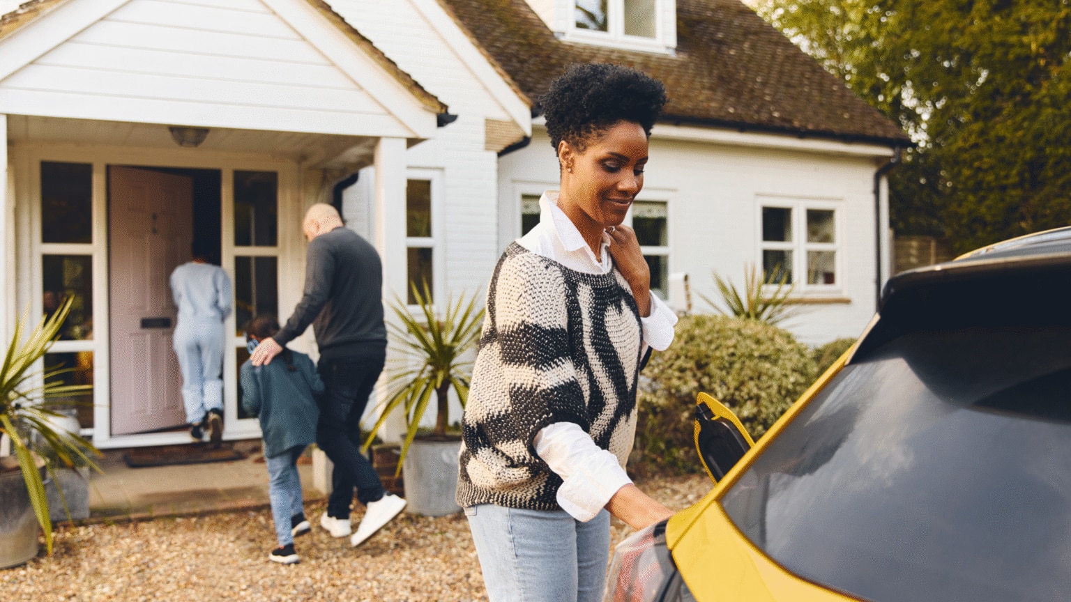 Children and man enters a house in background and woman standing by a yellow car in foreground
