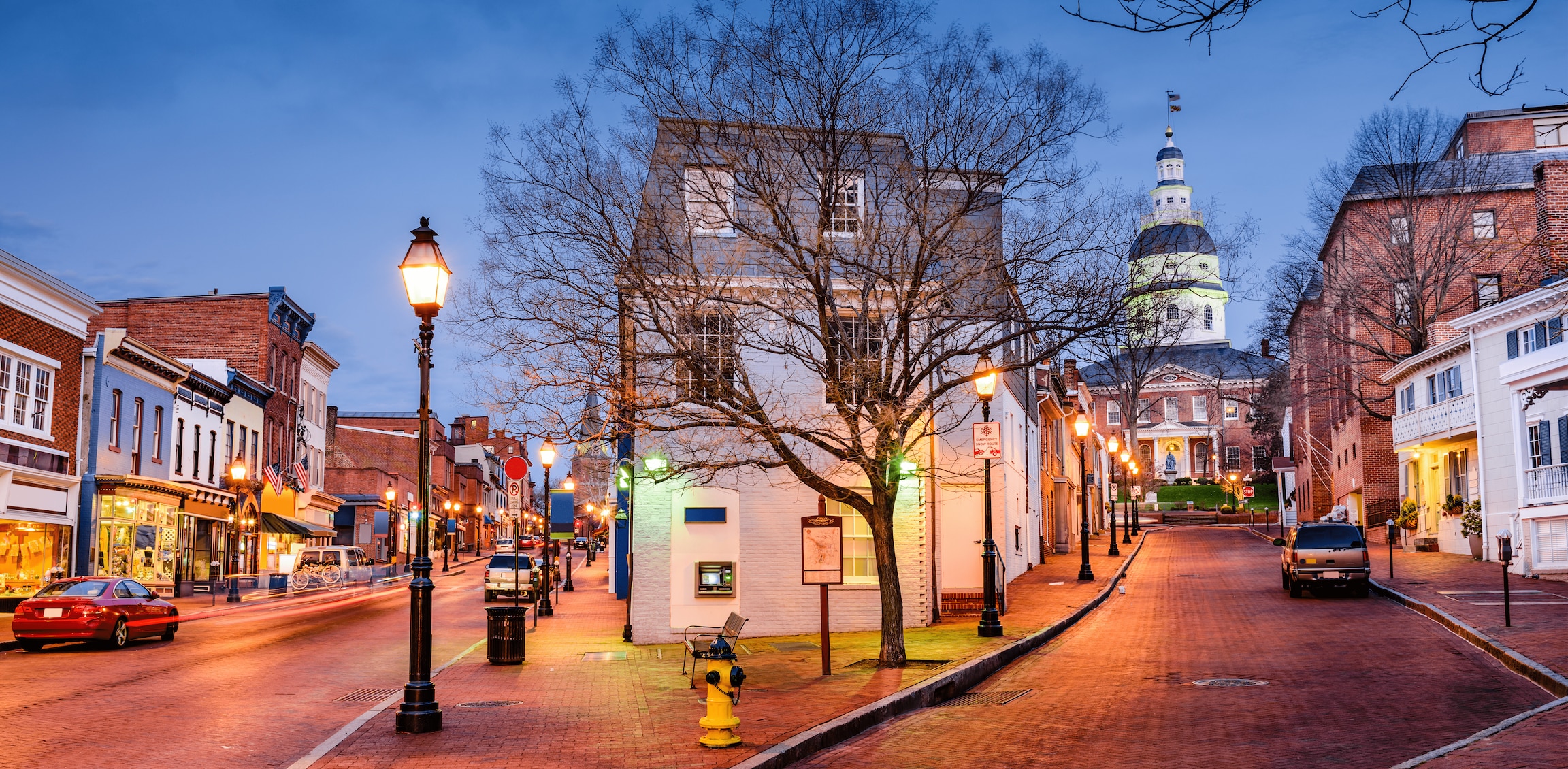 Historic downtown Annapolis, Maryland at dusk with lit storefronts and the Maryland State House in the background.