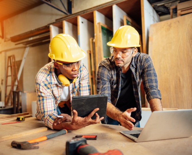 Two men wearing yellow hard hats standing at a desk with tools and a laptop on it.