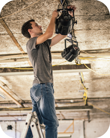 Man on a ladder adjusting hanging stage lights in an unfinished room with exposed ceiling wiring.