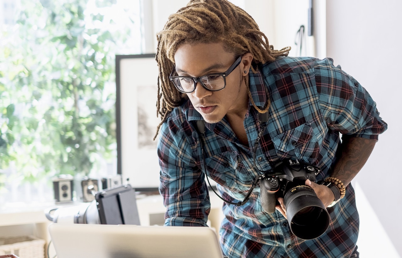 A photographer is holding a camera while looking at a laptop screen.