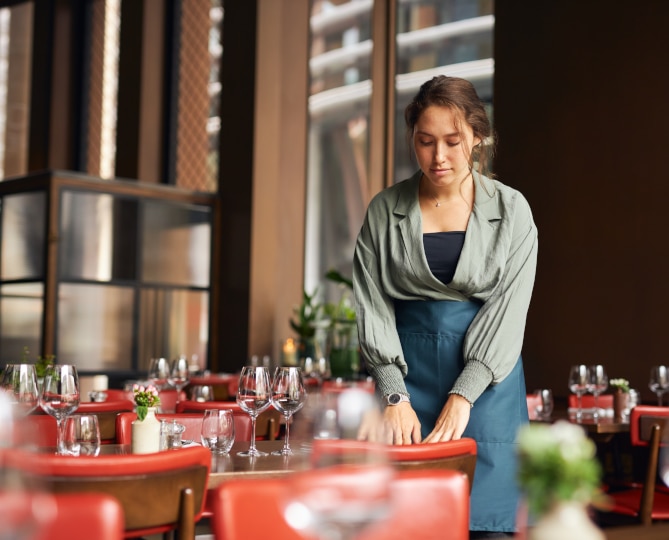 A woman wearing an apron setting a table inside a restaurant.