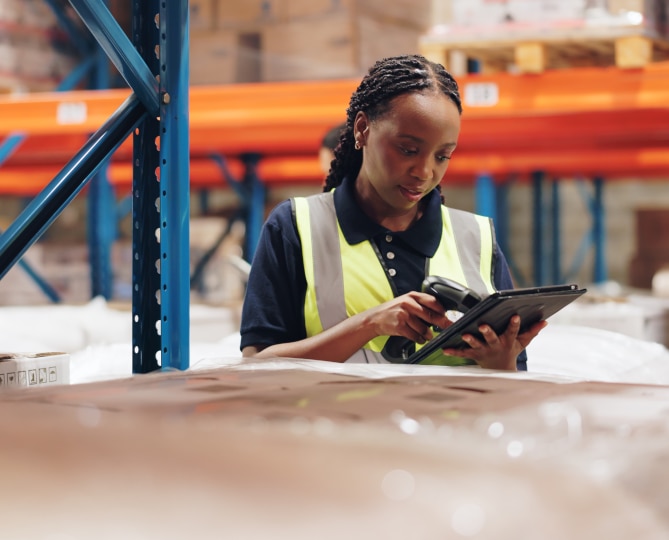 A Woman wearing a yellow reflective vest scanning boxes in a warehouse.