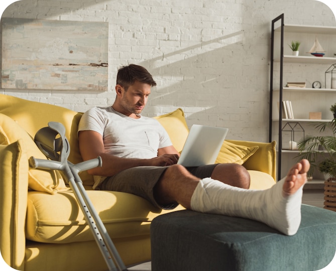 A man wearing a leg cast sitting on yellow couch looking at a laptop computer.