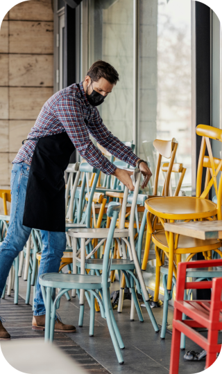 A man wearing an apron and mask stacking colorful chairs outside a restaurant.