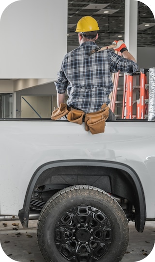 A Man with yellow hardhat and toolbelt sitting on the side of a truck bed.