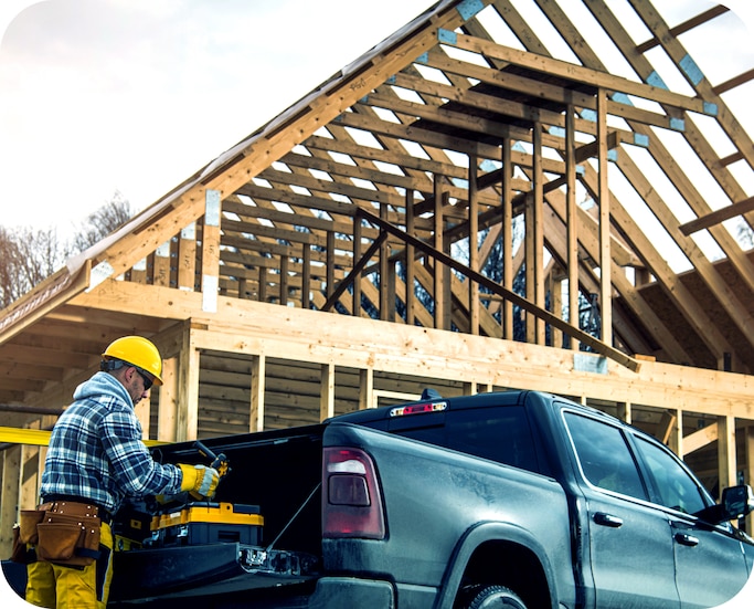 A man wearing a construction hat getting tools out of the bed of a black truck with a building under construction in the background.